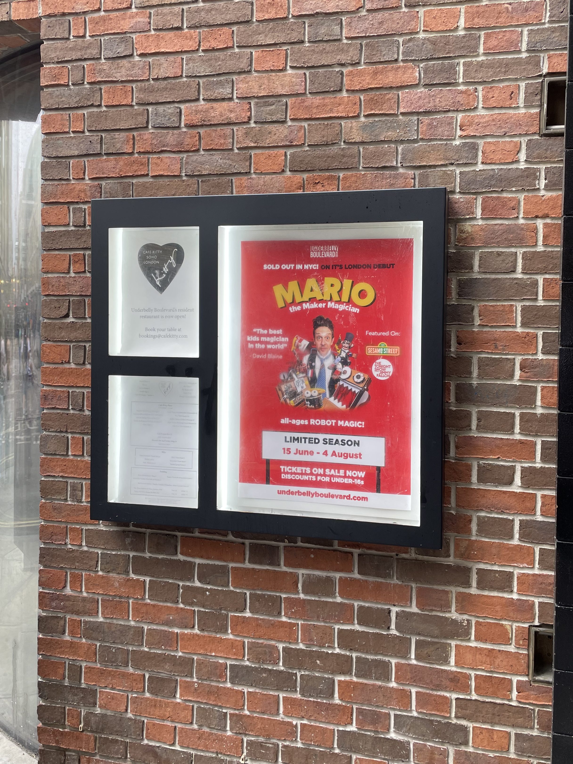 A poster box containing a red poster for Mario the Maker Magician on the brick wall of Underbelly Boulevard.